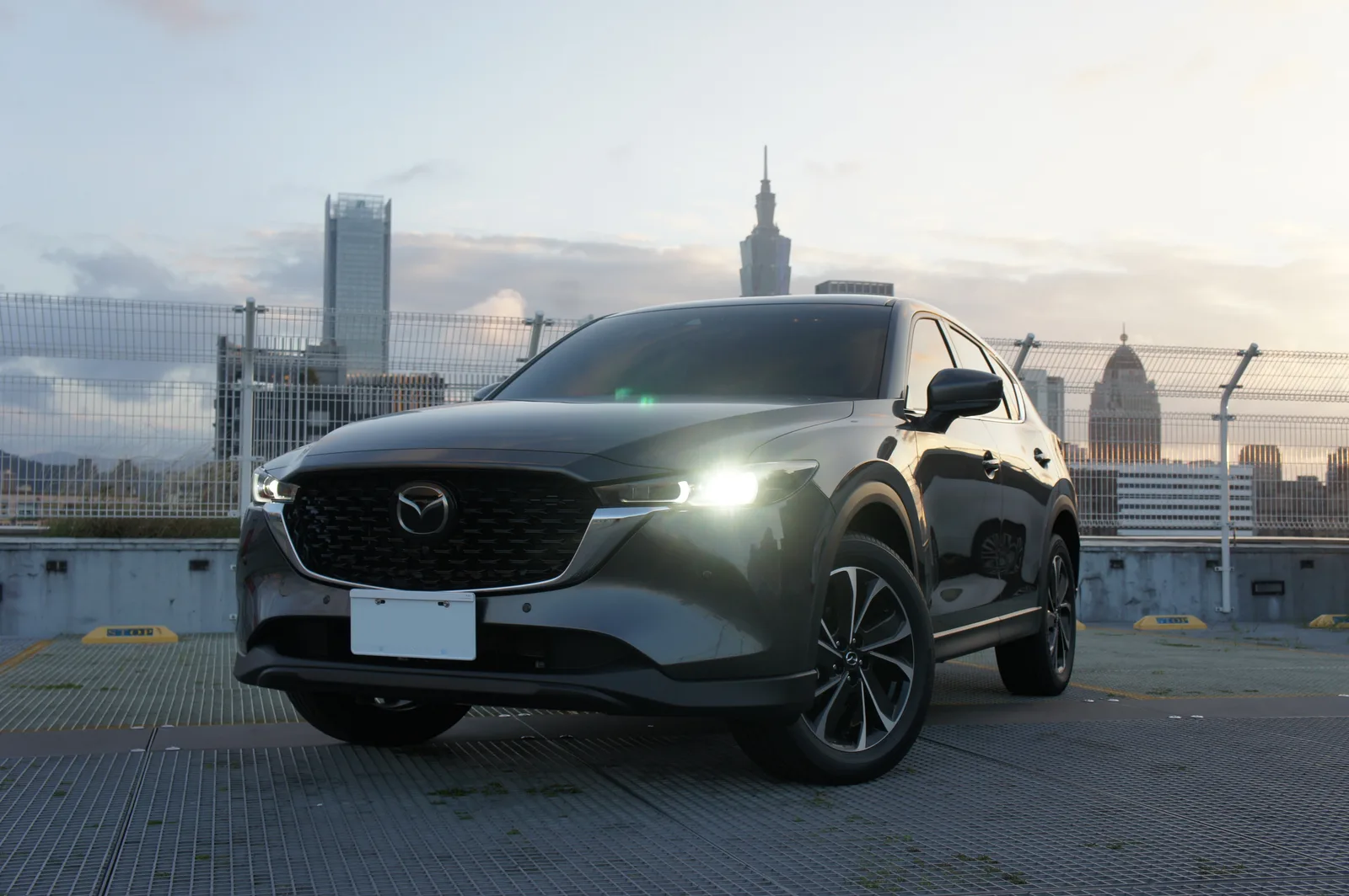 Mazda CX-5 2026 vue 3/4 avant en milieu urbain de nuit, coloris gris machine, finition Takumi avec feux laser