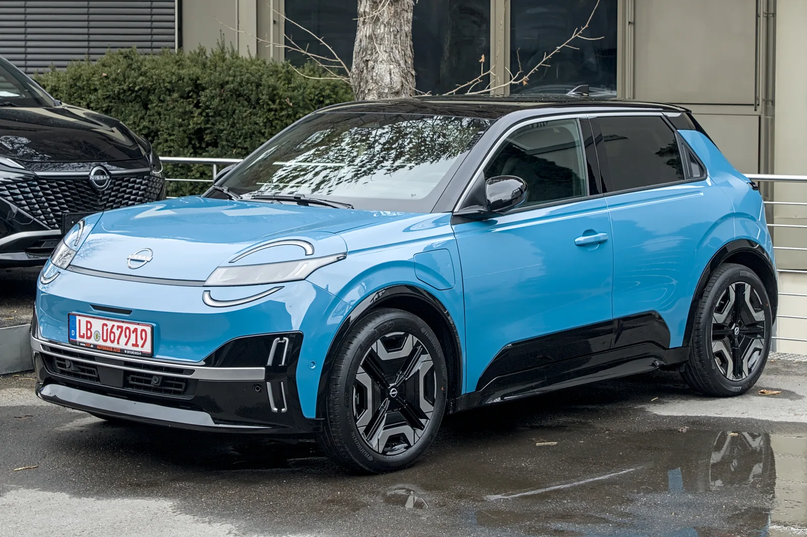 Nissan Micra électrique 2026 vue 3/4 avant garée devant un bâtiment, citadine bleue et noire