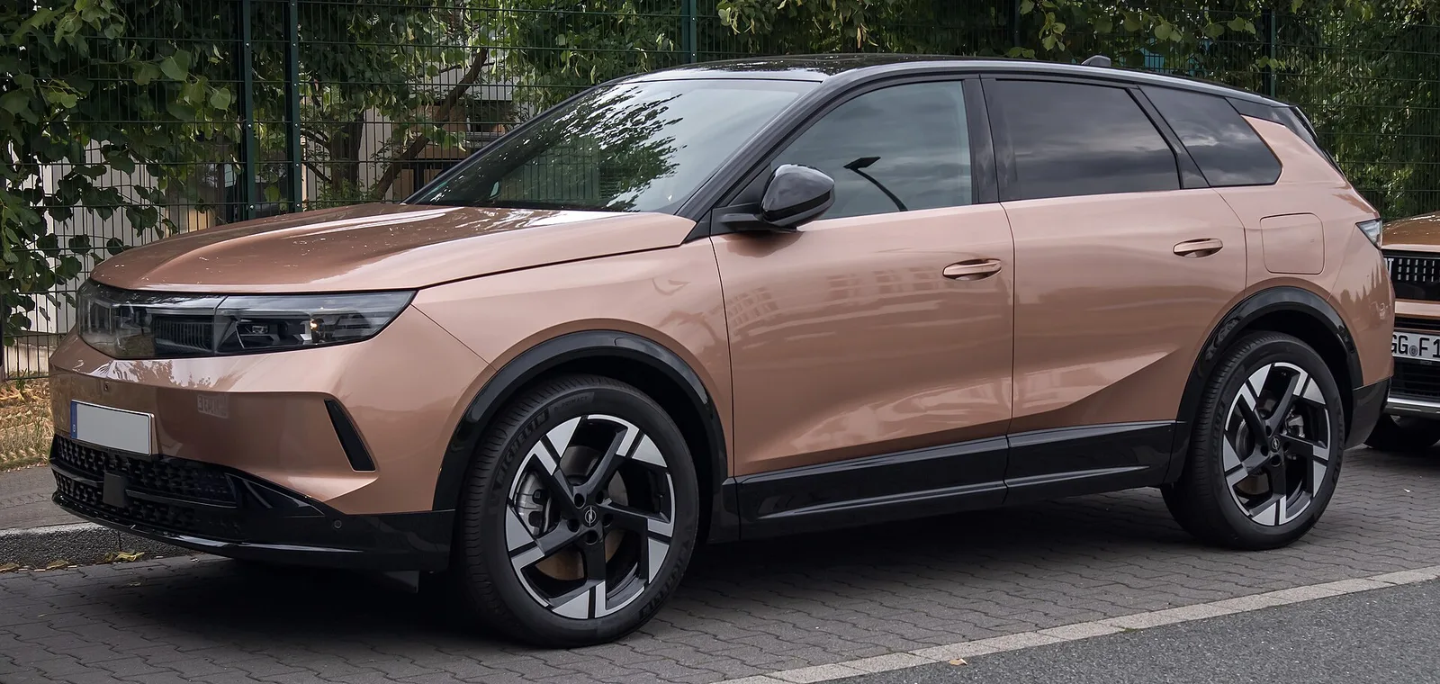 Opel Grandland Electric cuivré vu de trois quarts avant garé le long d'une grille, SUV compact électrique à 694 km WLTP en version Long Range