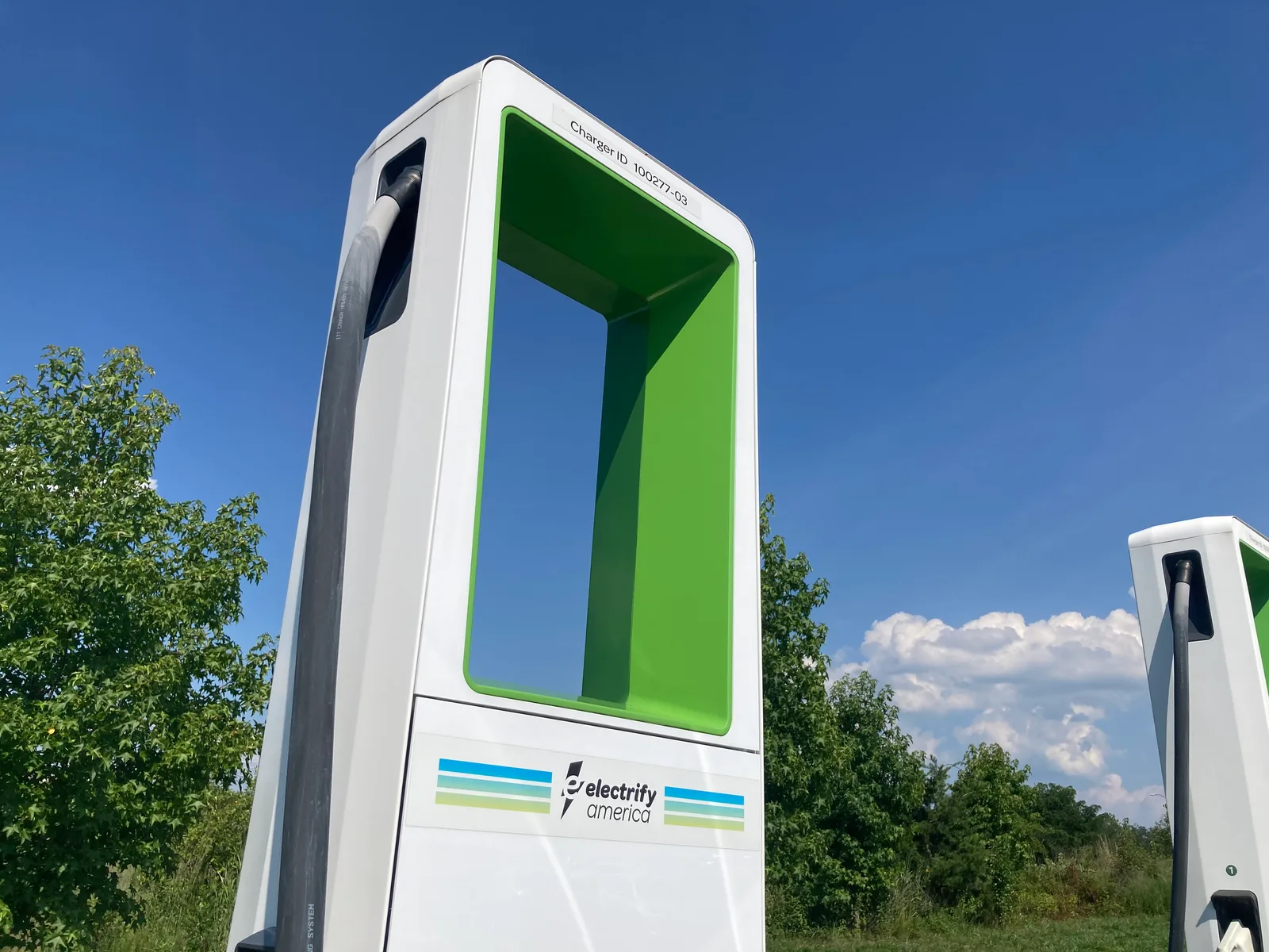 Borne de recharge rapide DC blanche et verte en extérieur, câble de charge suspendu, ciel bleu et végétation en arrière-plan