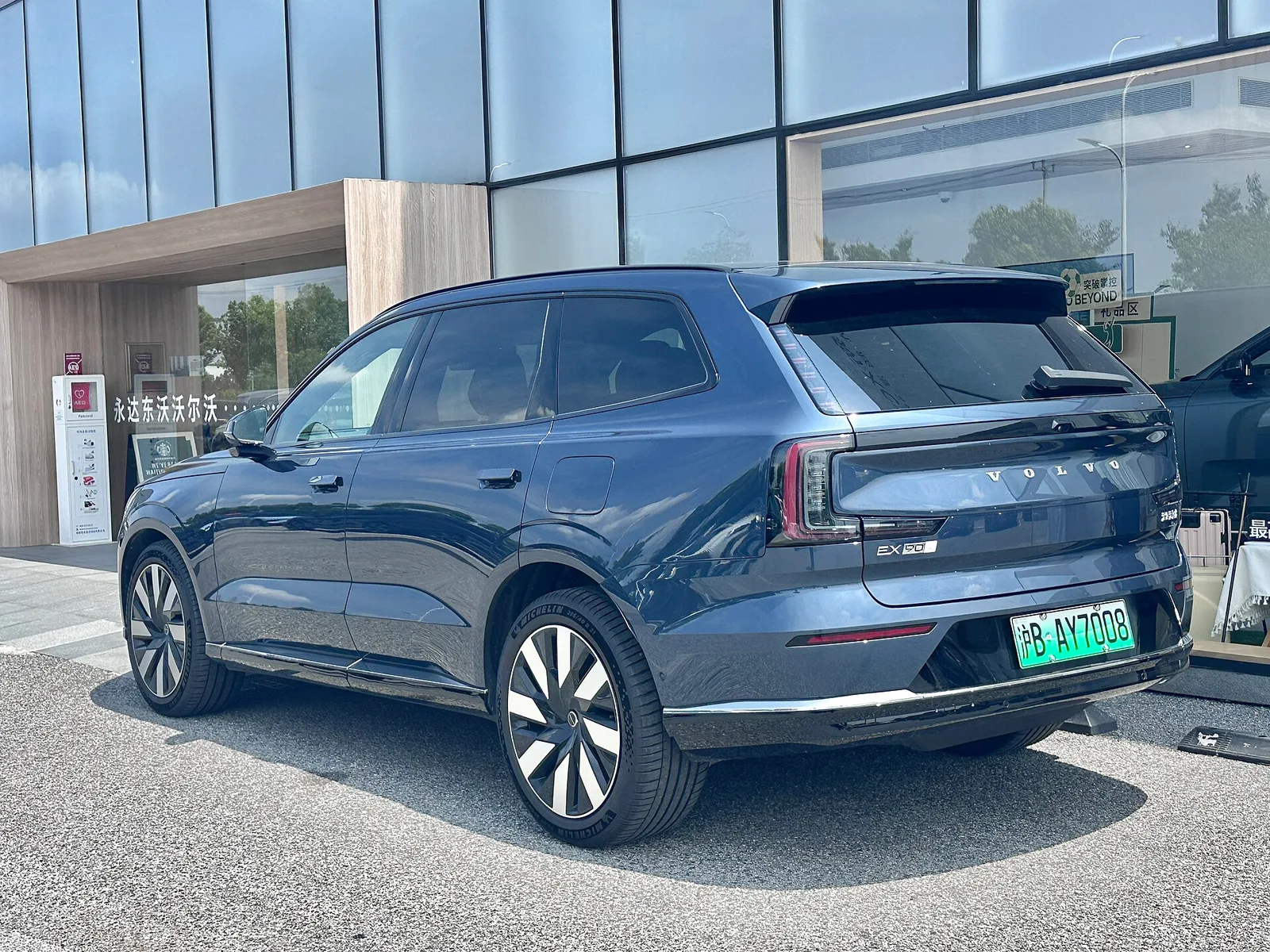 Volvo EX90 millésime 2026 bleu vu de trois quarts arrière devant une concession, SUV électrique 7 places à architecture 800 volts